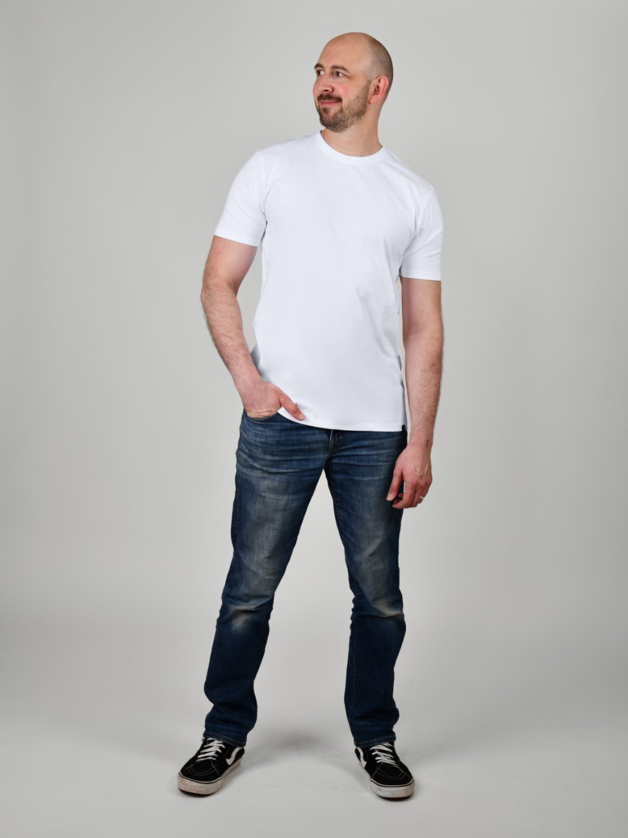 A head to toe shot of a tall and slim guy, hand in pocket, in the studio wearing a white XL tall slim t-shirt.