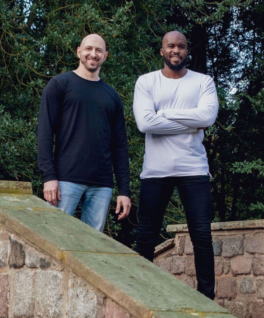 Two tall slim men wearing XL tall t-shirts, standing outdoors on a stone bridge and greenery in the background.