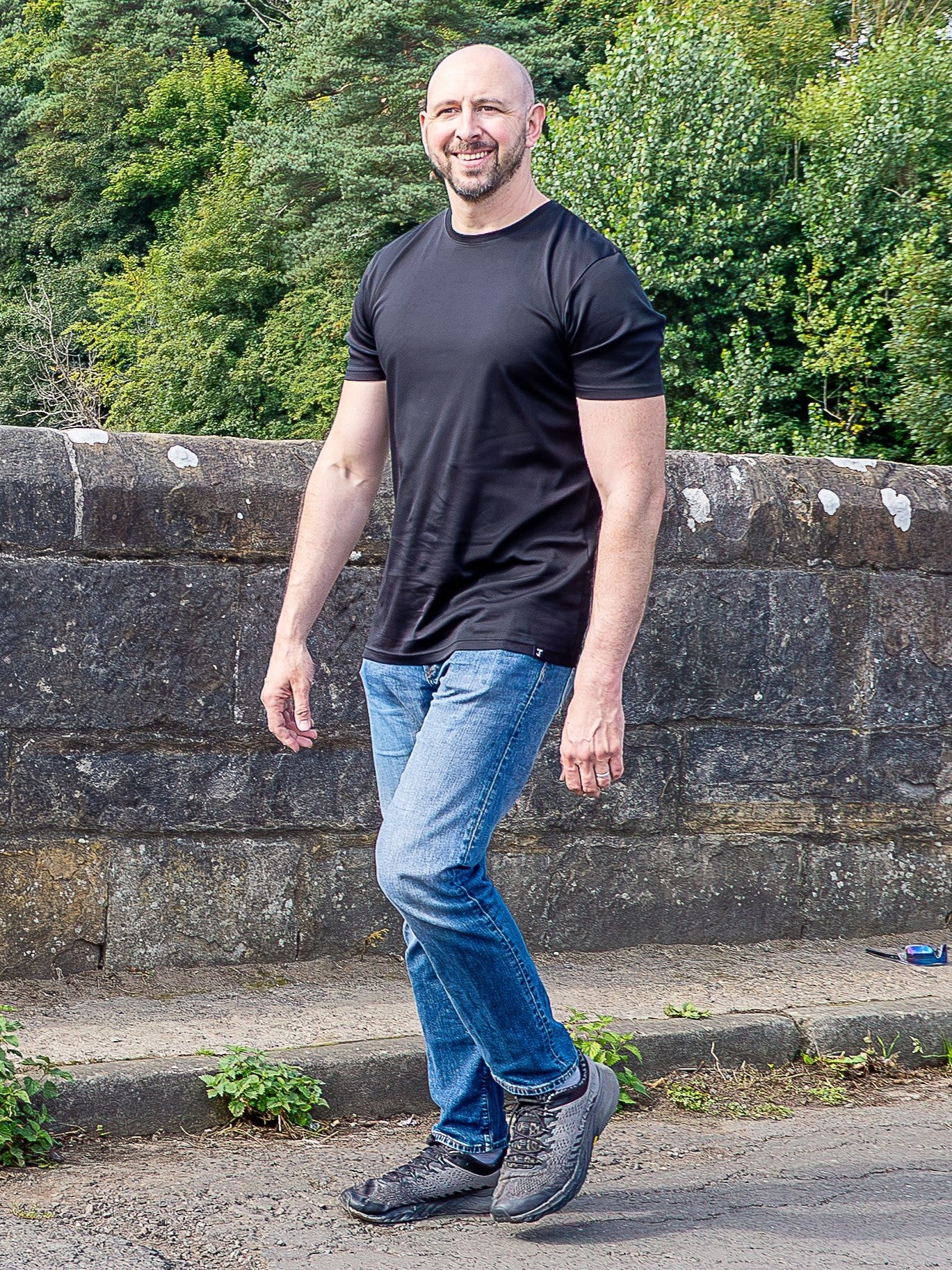 A man wearing a black tall mercerised t-shirt and blue jeans walking along a paved path next to a stone wall with a forest background.