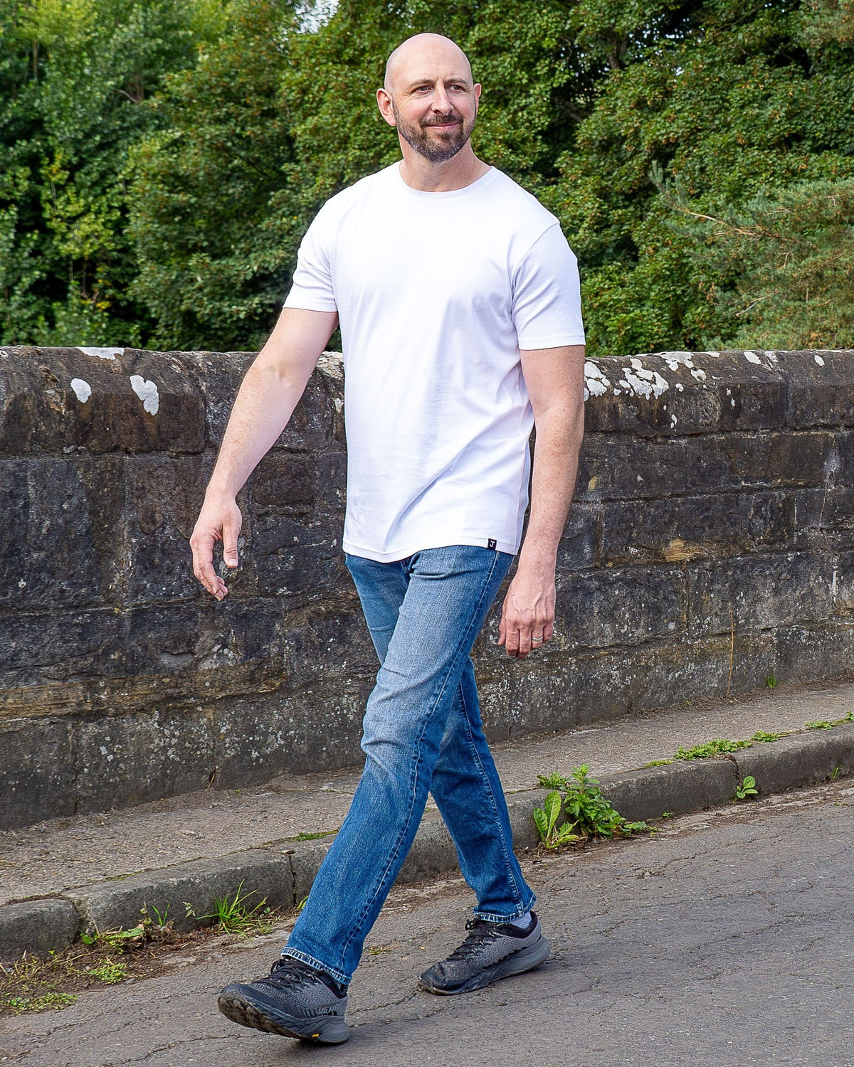 A man walking outdoors on a paved path wearing a white tall crew-neck t-shirt, blue jeans, and dark gray sneakers, with a stone wall and green trees in the background.